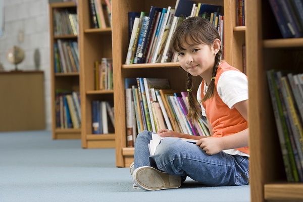 Girl (10-12) sitting on floor beside bookshelf in library, reading book, smiling